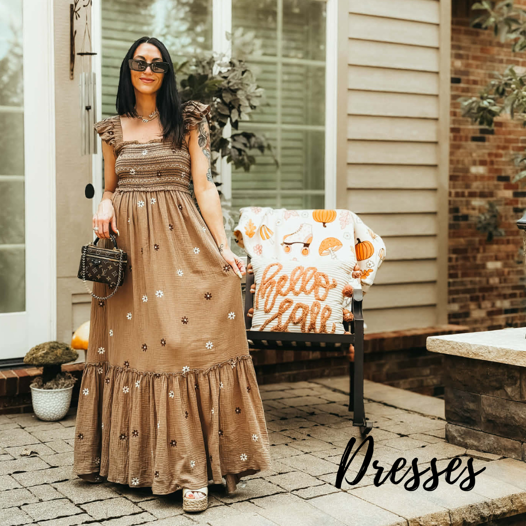 A woman stands outdoors on a patio wearing a long brown maxi dress with ruffled sleeves, embroidered floral details, and a tiered hem. She is holding a small patterned handbag, with a chair draped in a blanket behind her.